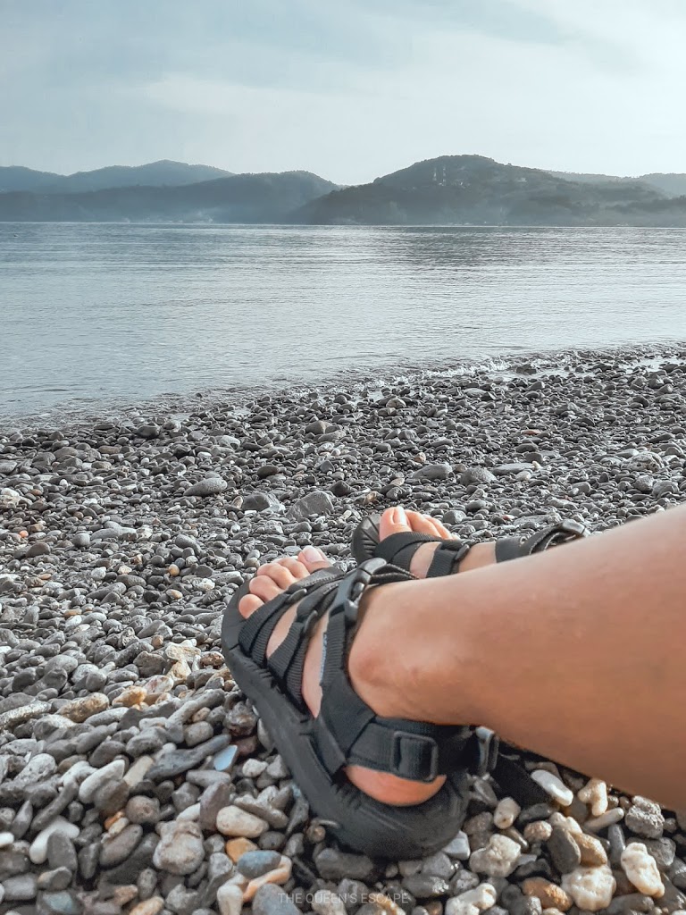 feet wearing a pair of black trekking sandals on a pebble beach